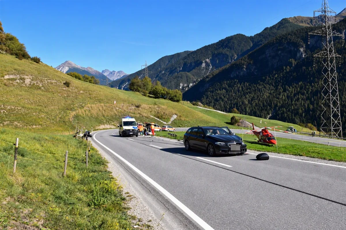 Ein Rettungshubschrauber ist auf einer Wiese neben einer Straße in einem Alpengebiet gelandet. Mehrere Einsatzfahrzeuge, darunter ein Krankenwagen, stehen auf der Straße, um bei einem Motorradunfall zu helfen.