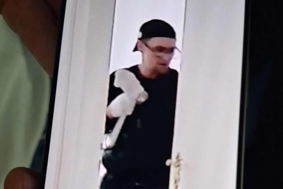 Ein Mann mit schwarzer Kappe und Brille steht in einer Türöffnung und hält einen weißen Gegenstand.