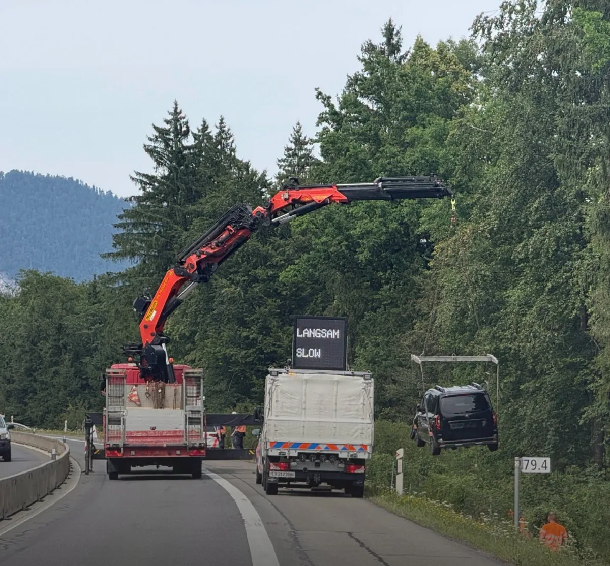 Ein schwarzes Auto wird von einem Kran neben einer stark befahrenen Autobahn angehoben. Ein Schild warnt mit 'Langsam'.