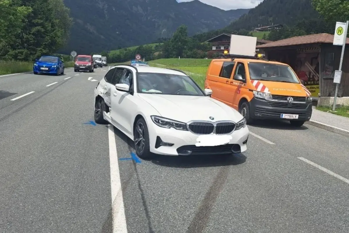 Ein weißer BMW mit sichtbaren Schäden an der Seite steht auf einer Straße. Daneben ist ein orangefarbener Van geparkt.