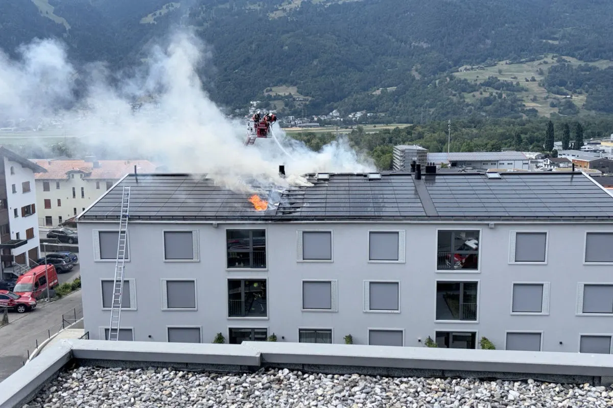 Auf dem Bild sieht man ein Mehrfamilienhaus, dessen Dach raucht und teilweise in Flammen steht. Ein Feuerwehrfahrzeug steht bereit, um den Brand zu löschen, während eine Leiter gegen das Gebäude lehnt.