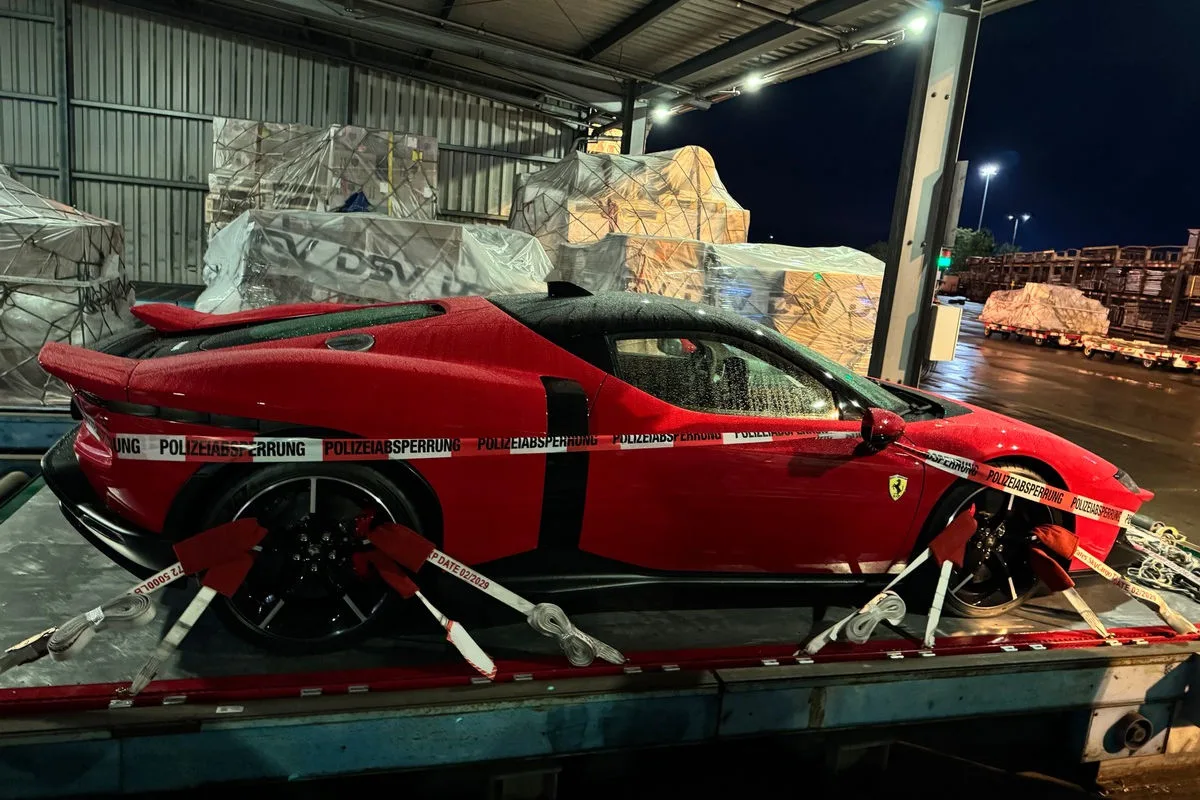 Ein roter Ferrari steht mit Polizeiabsperrband umwickelt auf einem Anhänger in einer Lagerhalle. Im Hintergrund sind Paletten zu sehen.