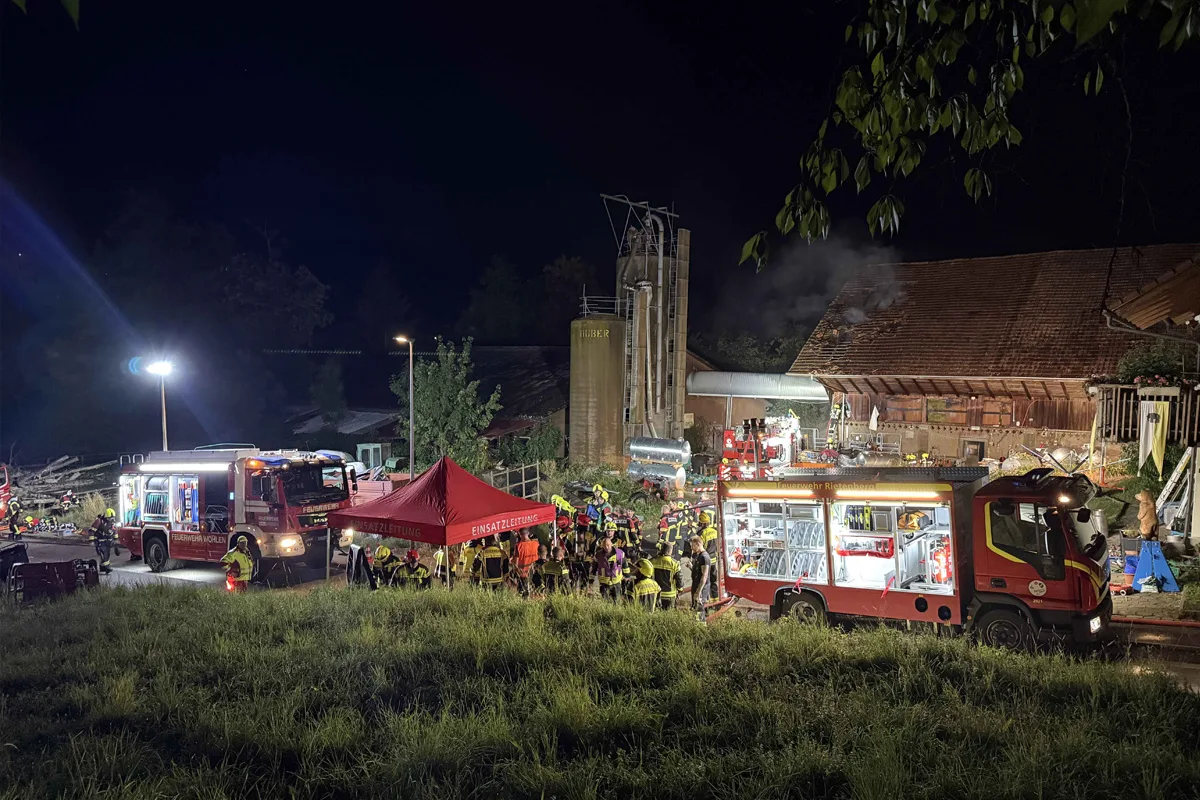 Zwei Feuerwehrfahrzeuge vor einem brennenden landwirtschaftlichen Gebäude mit Einsatzkräften, die das Feuer bekämpfen.