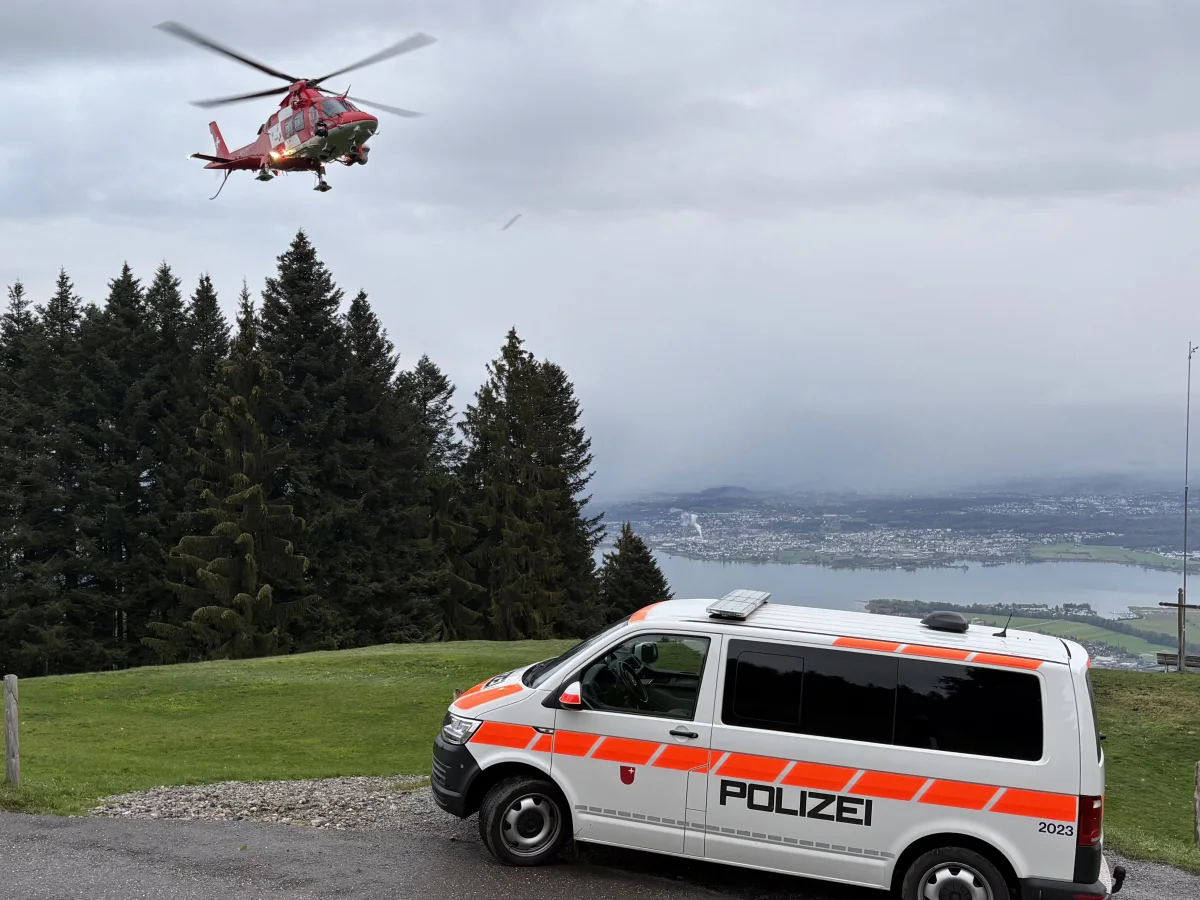 Ein weißer Polizeivan mit orangener Markierung steht auf einem Hügel, während ein roter Rettungshubschrauber in der Luft darüber schwebt, umgeben von Bäumen.