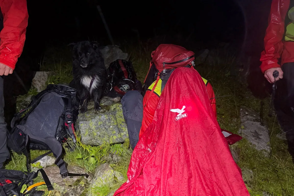 Mehrere Bergsteiger in wetterfester Kleidung sitzen bei Nacht auf felsigem Untergrund. Ein nasser Hund steht neben ihnen.
