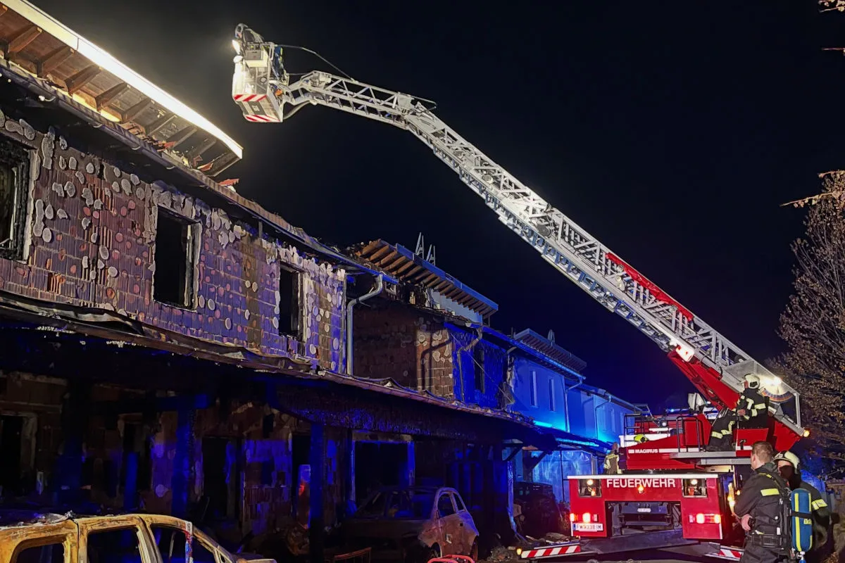 Ein Feuerwehrfahrzeug mit ausfahrbarer Leiter ist bei Nacht an einem teilweise ausgebrannten Haus im Einsatz. Das Gebäude zeigt deutliche Brandspuren und offene Fensteröffnungen.
