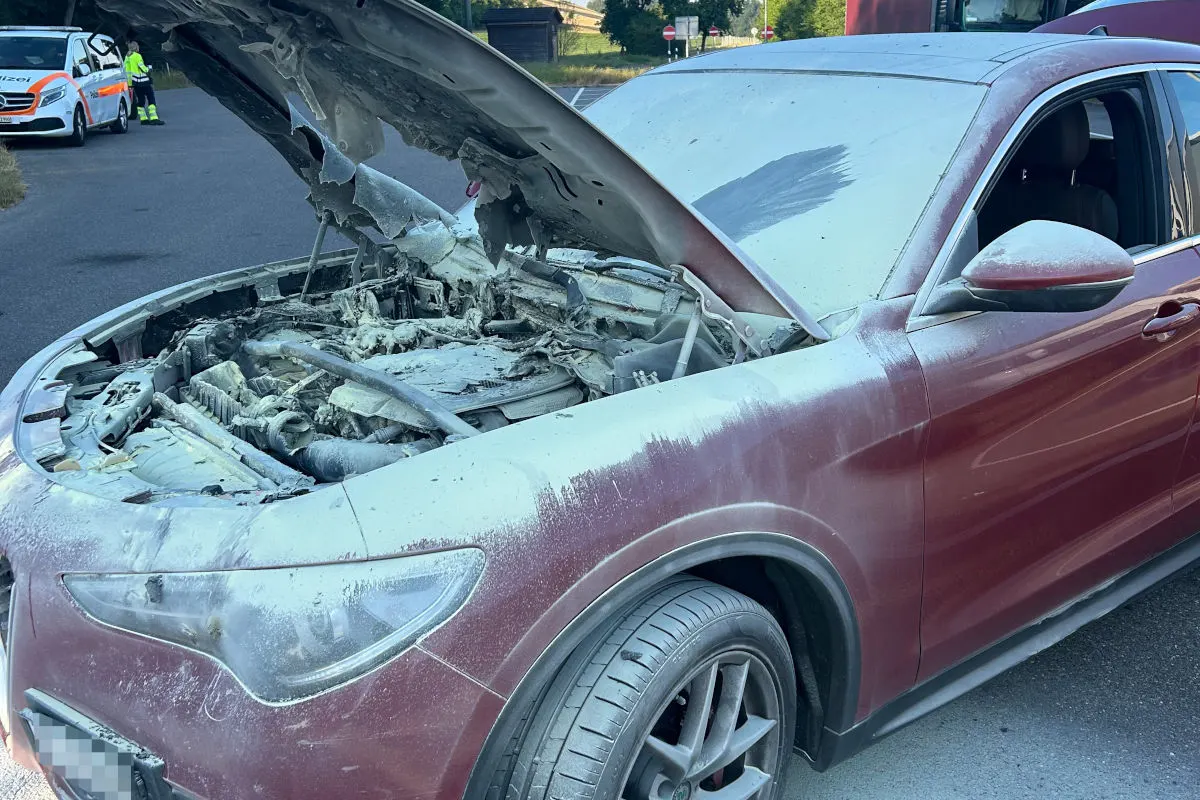 Ein Auto steht am Straßenrand mit offenem Motorraum, der ausgebrannt ist. Im Hintergrund ist ein Polizeifahrzeug sichtbar.
