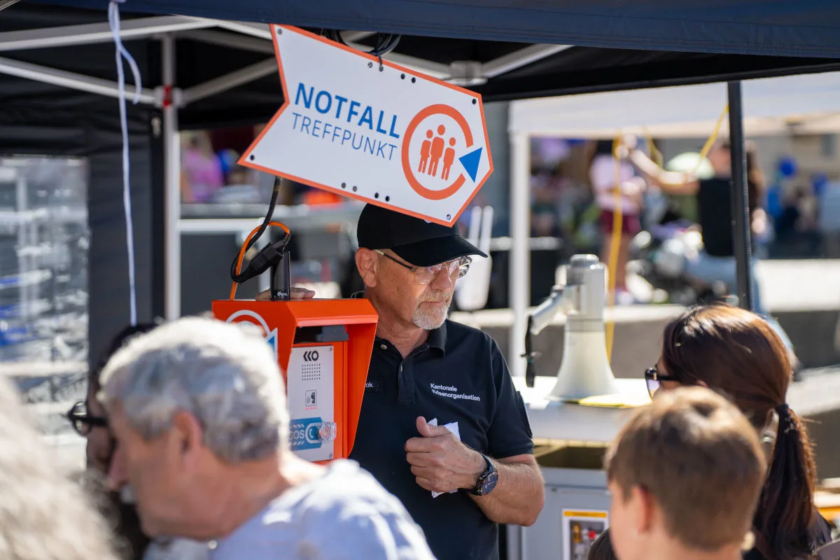 Ein älterer Mann trägt ein schwarzes Poloshirt mit der Aufschrift 'Kantonale Führungsorganisation' und steht neben einem orangefarbenen Gerät mit einem 'Notfall Treffpunkt'-Schild. Im Hintergrund sind Menschen und Zelte zu sehen.