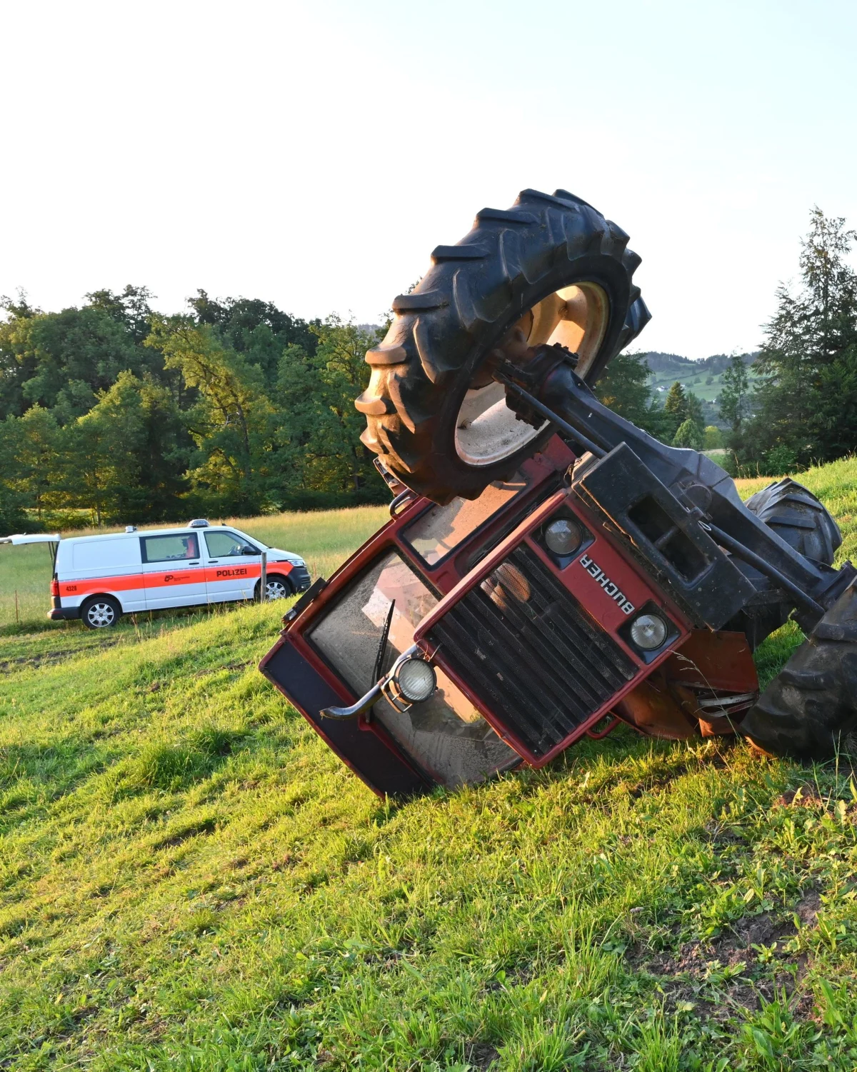 Ein roter Traktor liegt auf der Seite in einer grünen Wiesenlandschaft. Im Hintergrund ist ein Polizeifahrzeug zu sehen.