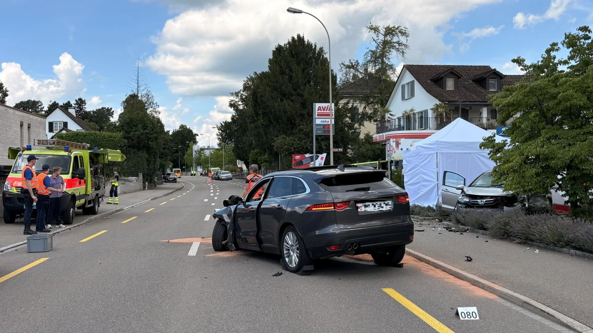 Ein graues Auto mit erheblichen Schäden steht auf einer Straße. Im Hintergrund ist eine Tankstelle und ein weiteres involviertes Auto zu sehen.