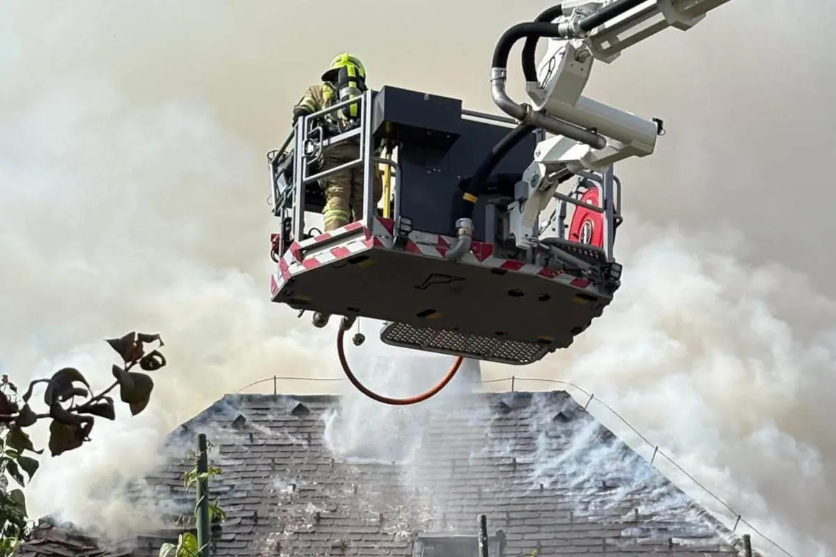 Ein Feuerwehrmann in voller Montur steht auf einer Hebebühne und bekämpft ein Feuer auf einem Dach, aus dem Rauch aufsteigt.