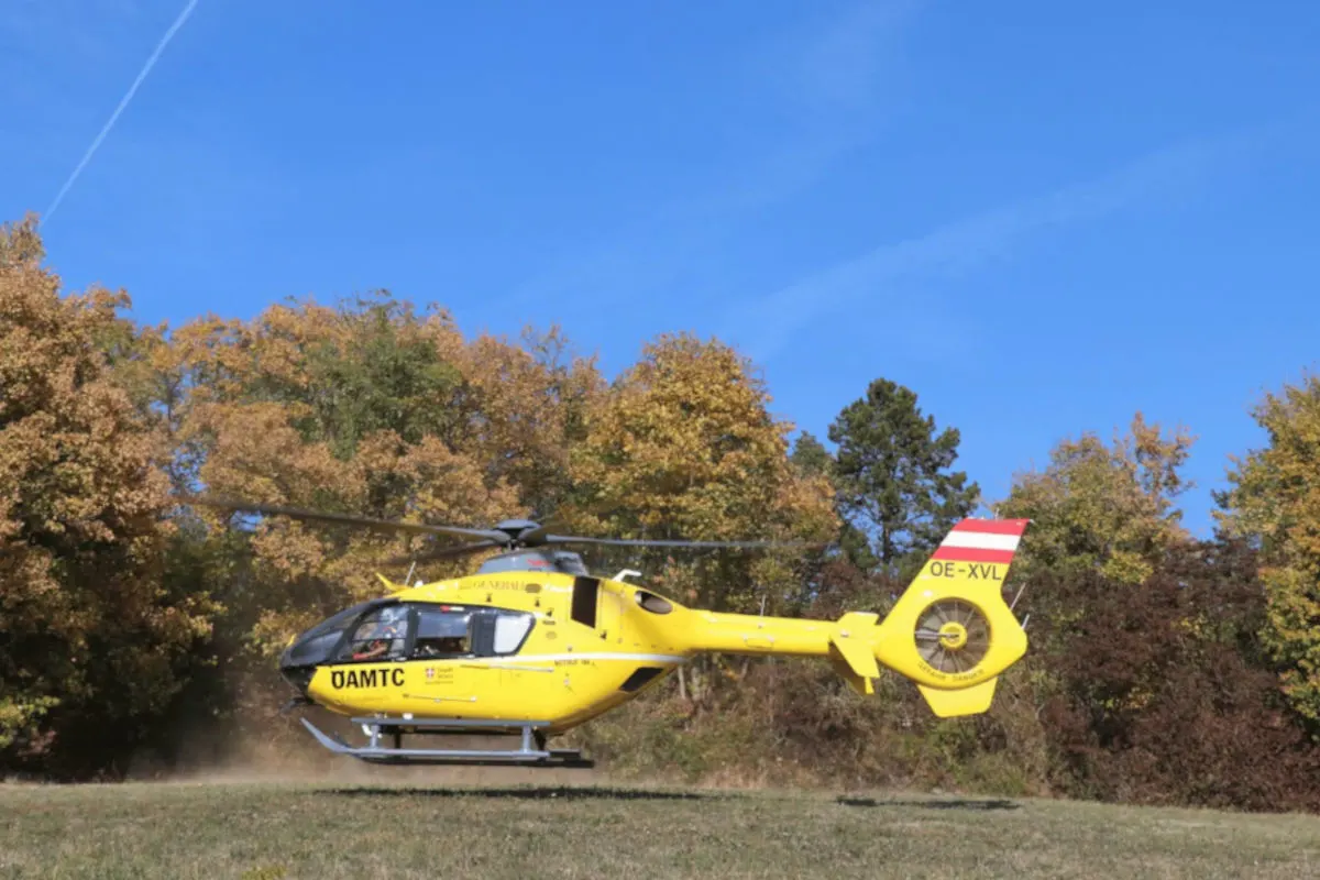 Ein gelber Rettungshubschrauber des ÖAMTC hebt von einer grasbewachsenen Fläche ab. Im Hintergrund sind herbstliche Bäume sichtbar.