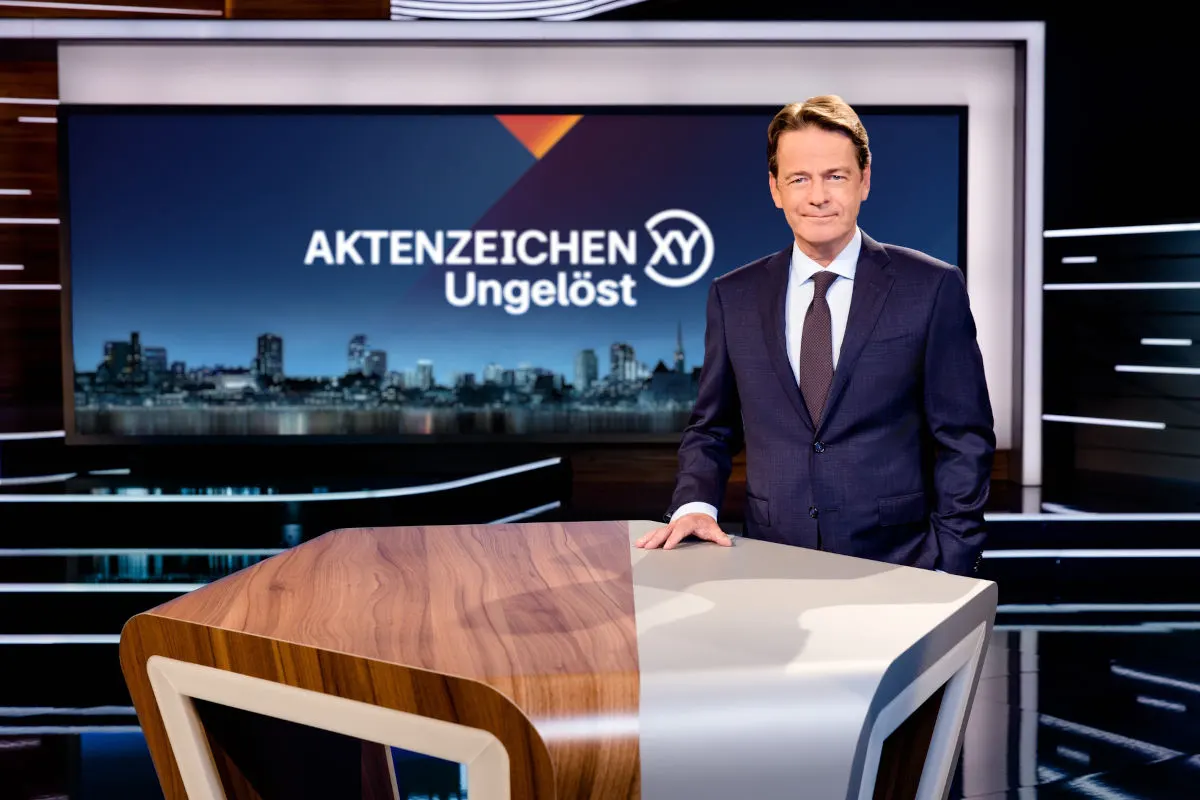 Ein Mann in einem dunklen Anzug steht vor einem hölzernen Tisch im Studio von Aktenzeichen XY ungelöst, mit einem Bildschirm im Hintergrund.