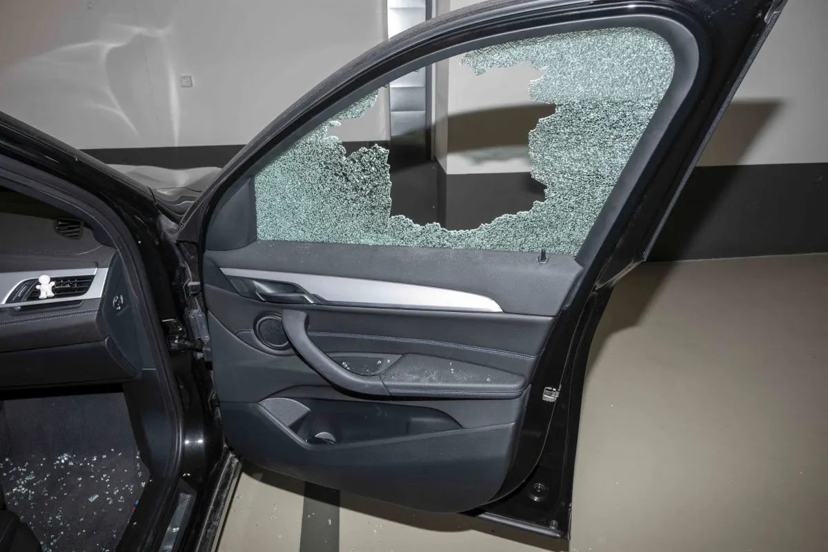 Die Seitenscheibe eines Autos ist eingeschlagen, wobei Glasstücke am Boden und auf dem Sitz verteilt sind. Die Tür des Fahrzeugs ist geöffnet und zeigt den Schaden deutlich.