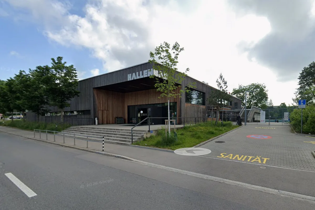 Ein modernes Gebäude mit der Aufschrift 'Hallenbad', umgeben von Bäumen. Der Himmel ist leicht bewölkt.