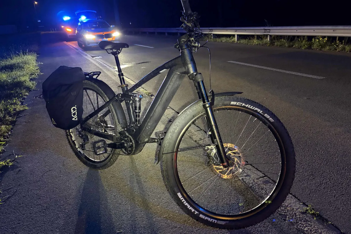 Ein E-Bike mit seitlicher Tasche ist nahe einer Fahrbahnkante positioniert. Im Hintergrund steht ein Polizeiauto mit eingeschaltetem Blaulicht.