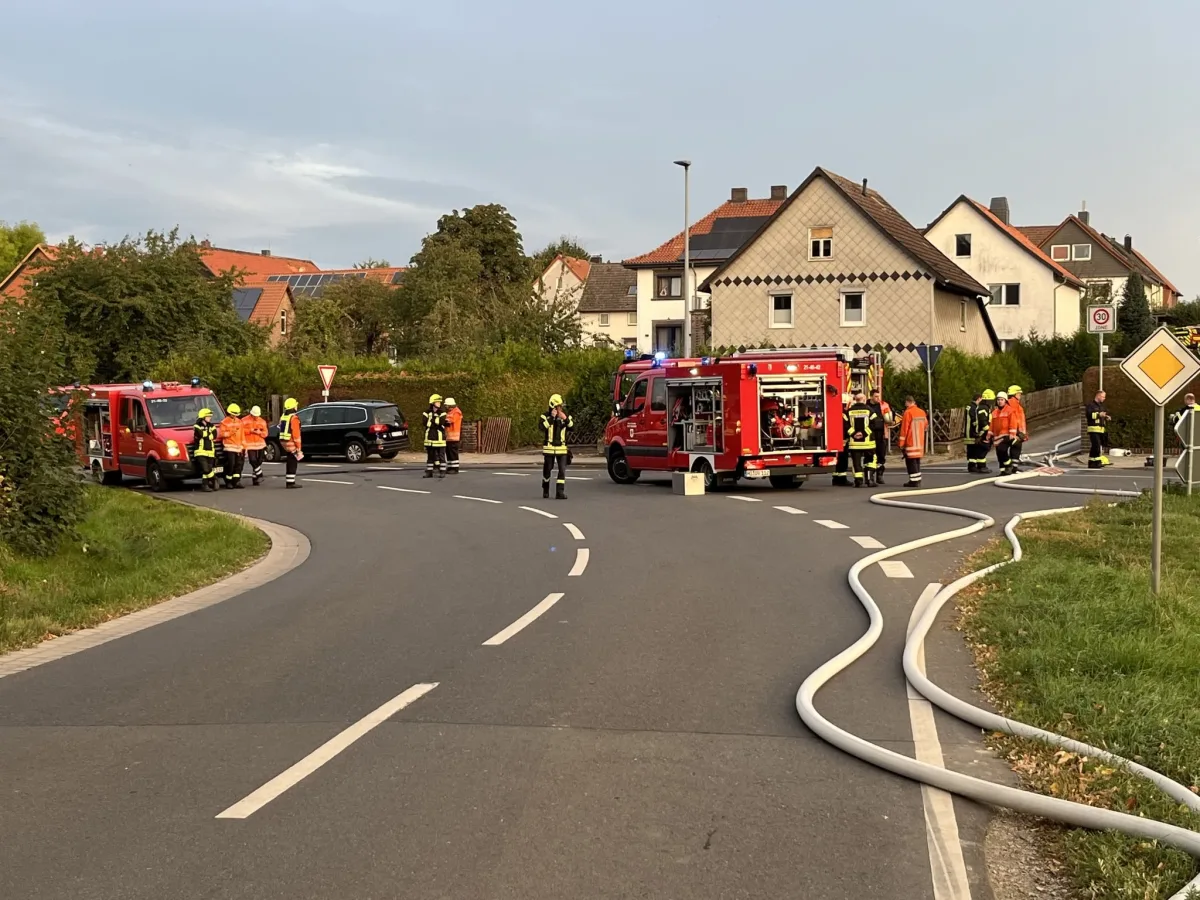 Mehrere Feuerwehrfahrzeuge und Feuerwehrleute sind an einer Straßeneinmündung in einem Wohngebiet im Einsatz. Schläuche liegen auf der Straße.