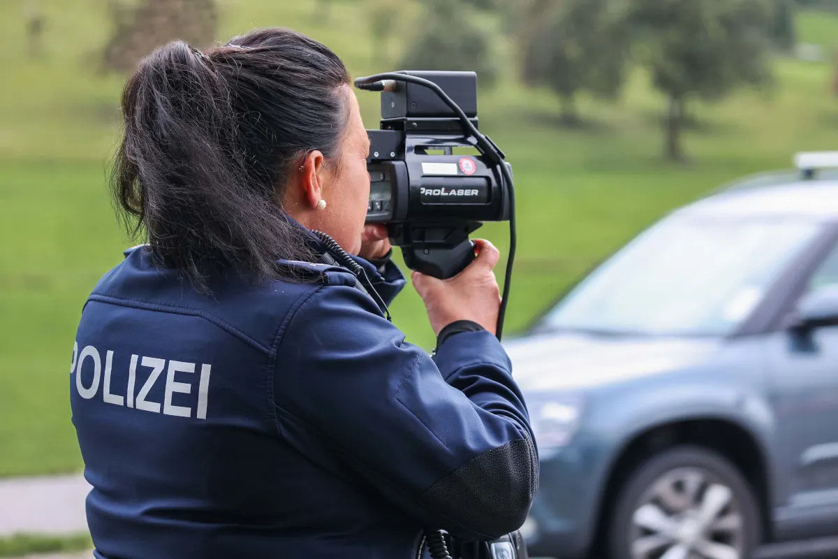 Eine Polizistin in dunkler Uniform hält ein Laser-Geschwindigkeitsmessgerät und zielt auf ein vorbeifahrendes Fahrzeug ab. Im Hintergrund ist ein Auto sichtbar.