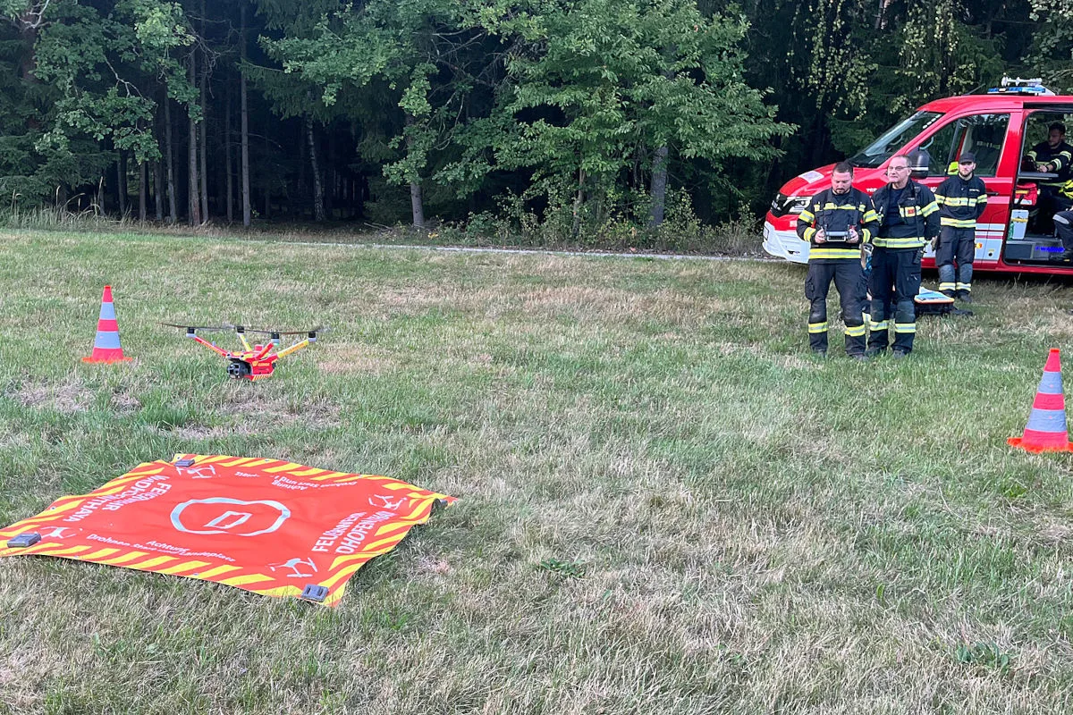Eine Drohne steht auf einer Markierung vor einem Feuerwehrfahrzeug mit drei Feuerwehrleuten auf einer Wiese nahe eines Waldes.