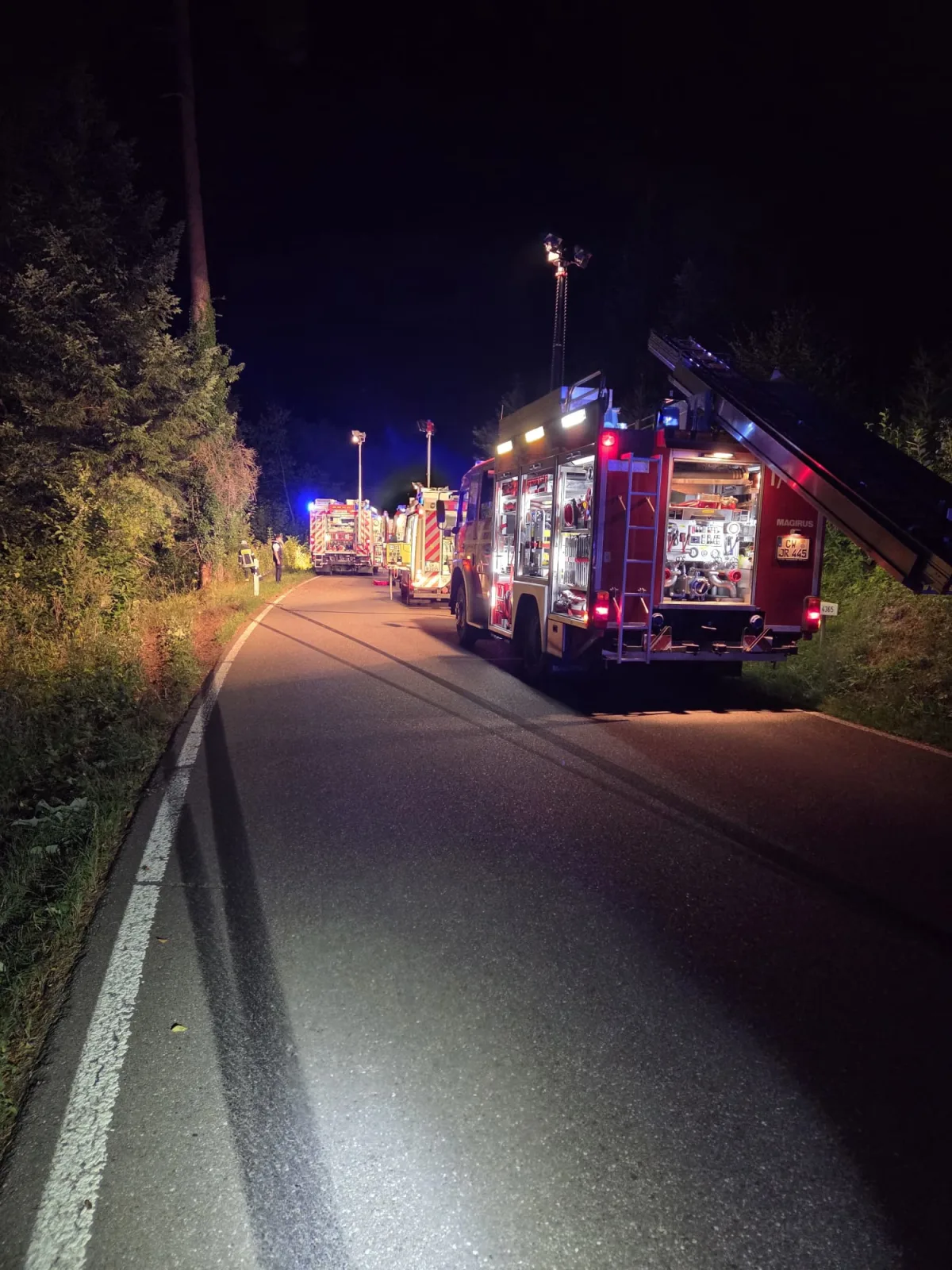 Mehrere Feuerwehrfahrzeuge parken auf einer Waldstraße bei Nacht, beleuchtet von ihren Lichtern. Der Bereich wird von der Feuerwehr abgesichert.