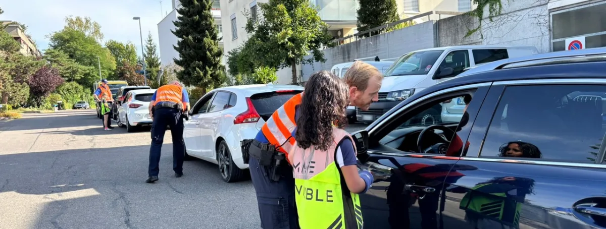 Zwei Polizisten in orangefarbenen Westen kontrollieren Autofahrer auf einer sonnigen Wohnstraße. Mehrere geparkte Autos sind zu sehen.