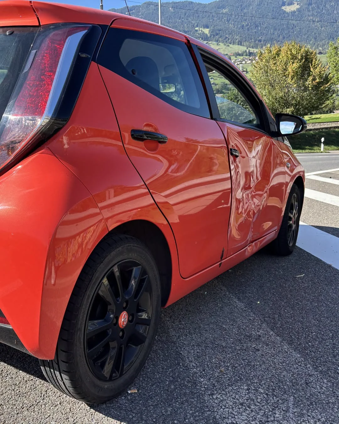 Ein rotes Auto steht auf der Straße mit einer deutlichen Delle an der Beifahrertür. Es ist tagsüber, bei klarem Wetter.