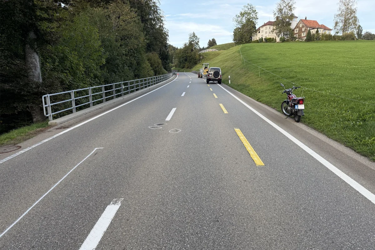 Eine Landstraße mit einem geparkten Auto am Rand und einem Motorrad auf dem Seitenstreifen neben einer Wiese.