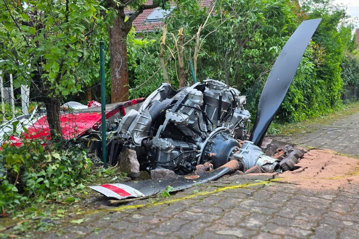 Ein abgestützter Flugzeugmotor liegt auf einem gepflasterten Gehweg zwischen Bäumen und Büschen. Trümmerteile verteilen sich um das Wrack.