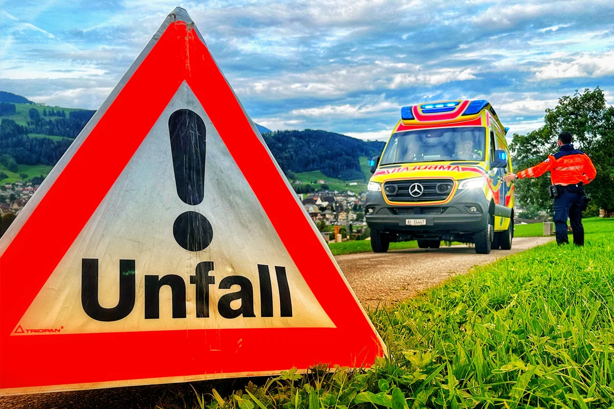 Ein dreieckiges Unfallwarnschild steht im Vordergrund auf einer Landstraße. Im Hintergrund ist ein buntes Ambulanzfahrzeug zu sehen, umgeben von grüner Landschaft und einem bewölkten Himmel.