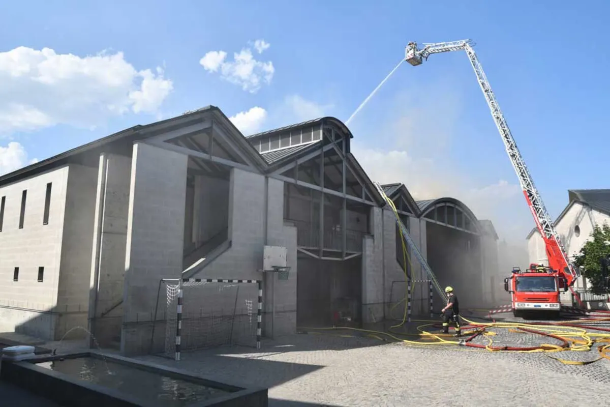 Ein Feuerwehrfahrzeug mit ausfahrbarer Leiter löscht Rauchschwaden von einem großen Industriegebäude, während Feuerwehrleute mit Schläuchen arbeiten.