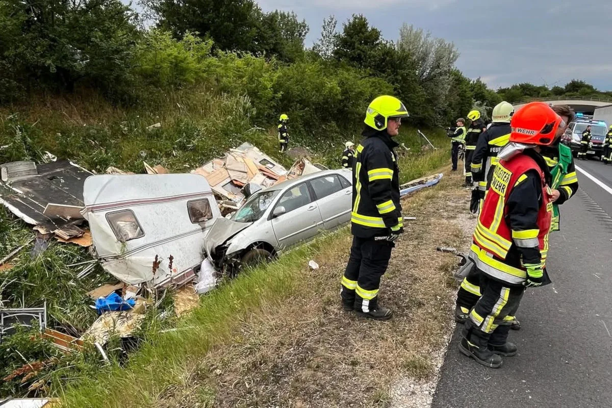 Ein verbeulter Pkw steht neben einem zerstörten Wohnwagen im Straßengraben. Feuerwehrkräfte sind vor Ort, um die Situation zu sichern.