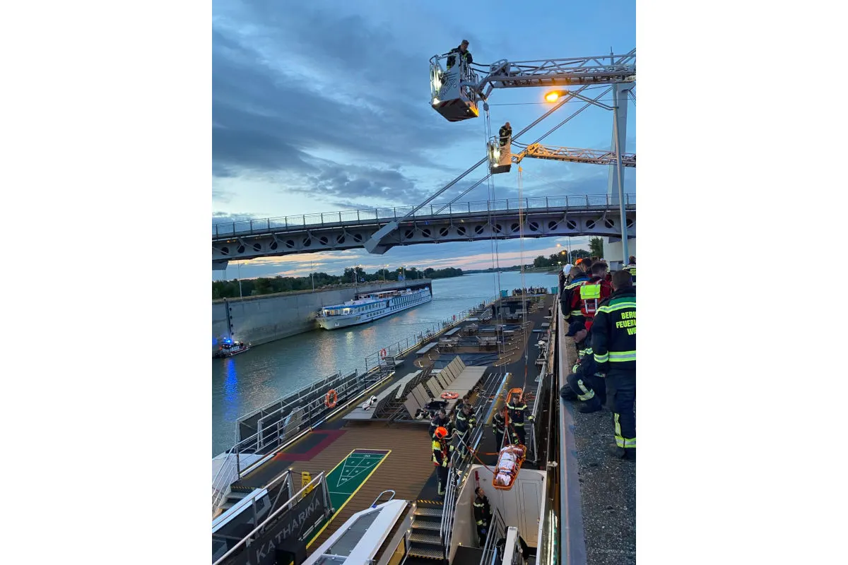 Die Feuerwehr nutzt einen Kran, um Ausrüstung auf das Deck eines unter einer Brücke liegenden Schiffs zu transportieren. Mehrere Einsatzkräfte stehen auf dem Schiffsdeck bereit.