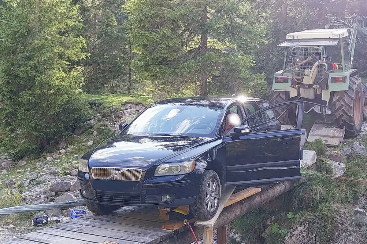Ein schwarzer Volvo befindet sich auf einer provisorischen Holzbrücke in einer bewaldeten Gegend. Im Hintergrund ist ein Traktor zu sehen.