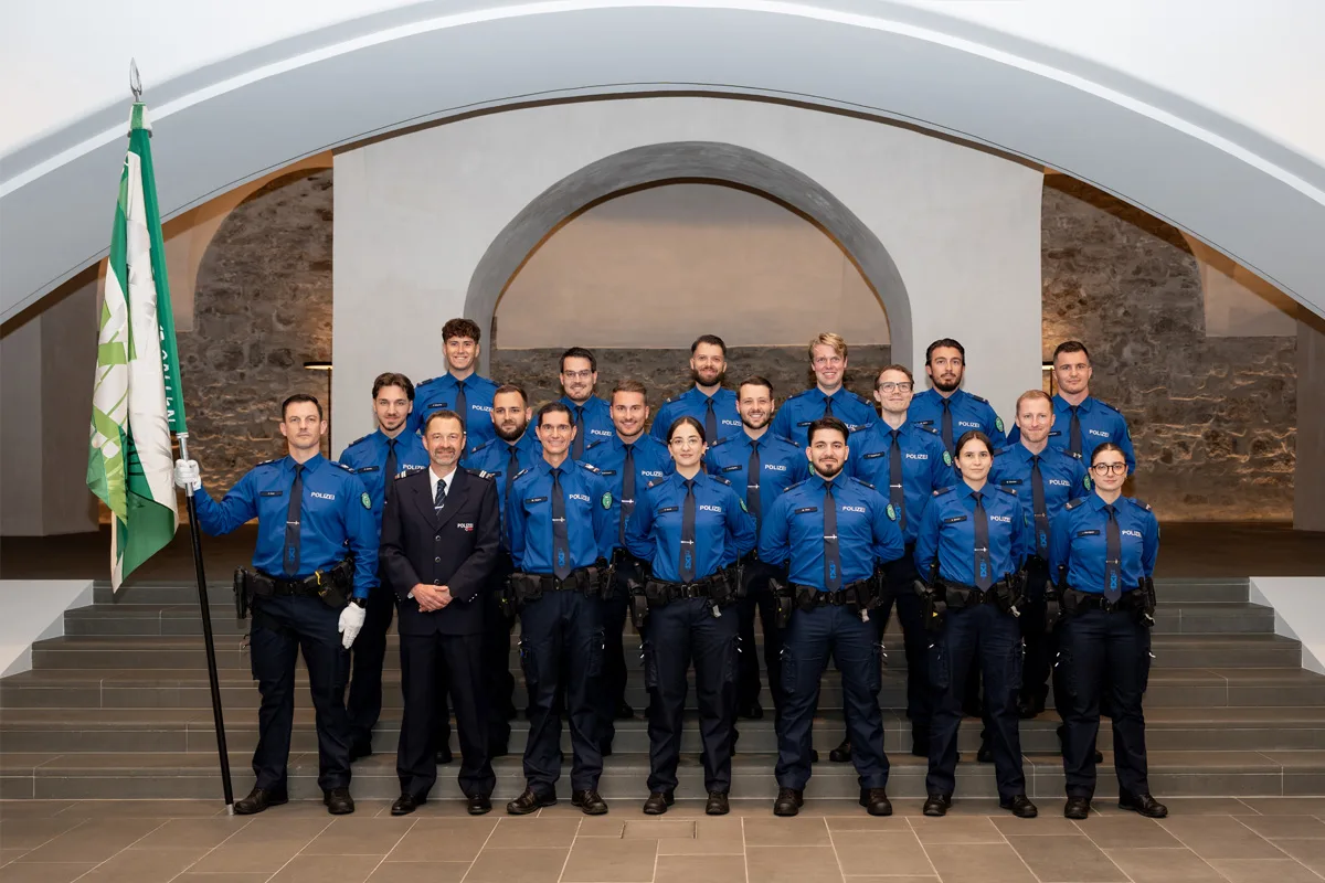 Eine Gruppe von etwa 20 Polizisten in blauen Uniformen steht auf Stufen in einem hellen Innenraum. Ein Polizist hält eine grüne Flagge.