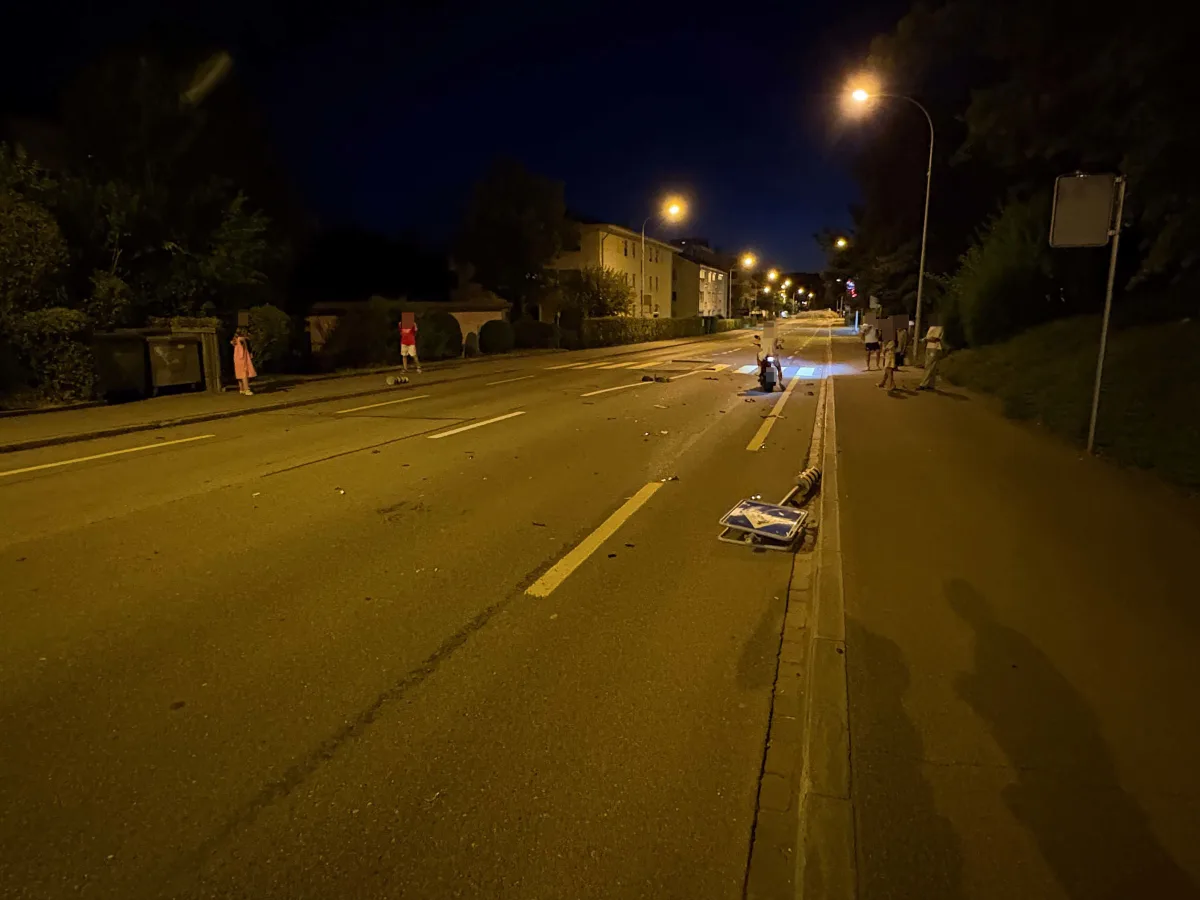 Eine nächtliche Straße in einer Wohngegend mit einem umgestürzten Verkehrsschild auf der Fahrbahn. Passanten beobachten die Szene vom Gehweg aus.