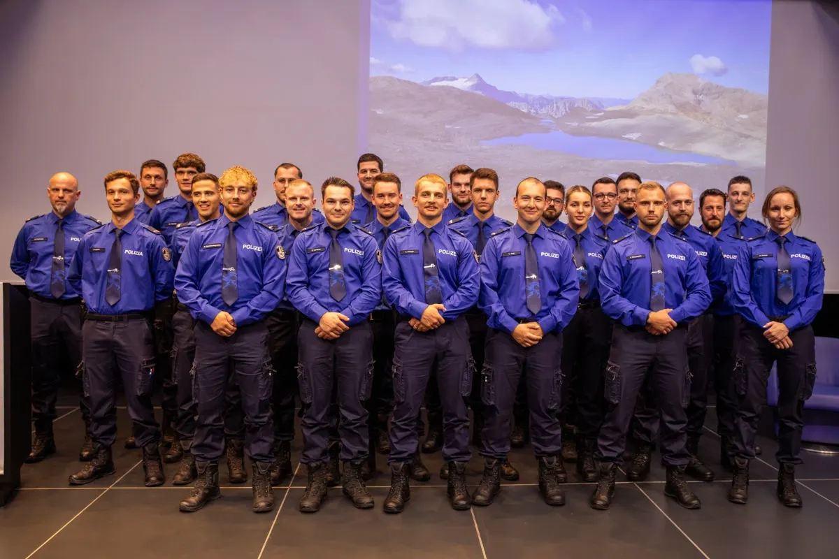 Eine Gruppe von Polizisten in blauer Uniform steht in mehreren Reihen bei einer feierlichen Veranstaltung. Hinter ihnen ist ein Bildschirm mit einem Bergpanorama sichtbar.