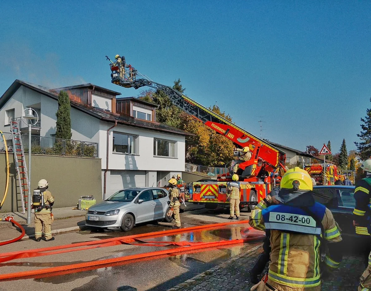 Mehrere Feuerwehrleute bekämpfen ein Feuer auf dem Dach eines zweistöckigen Hauses mit einer Drehleiter. Im Vordergrund stehen Feuerwehrfahrzeuge und Schläuche.
