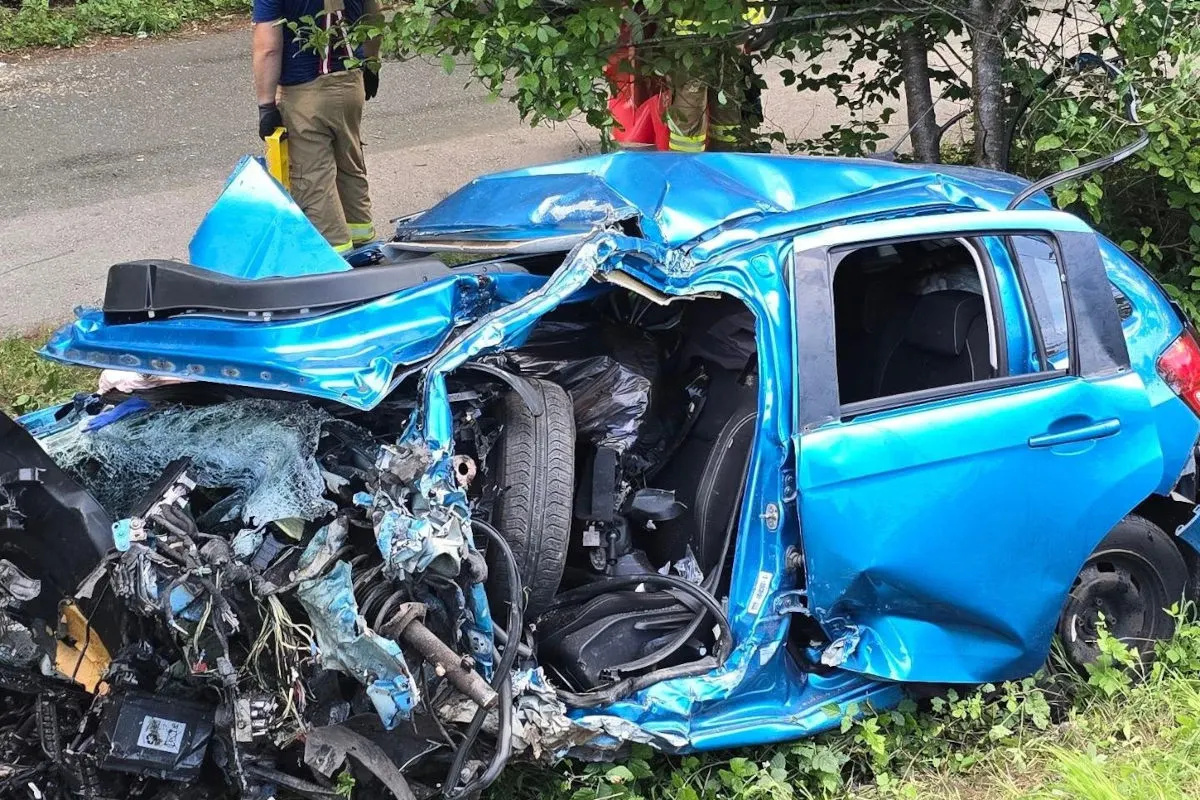 Ein stark verformtes blaues Auto steht am Straßenrand nach einem heftigen Zusammenstoß. Zwei Einsatzkräfte sind im Hintergrund zu sehen.