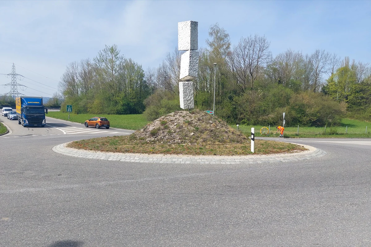 Im Vordergrund befindet sich eine große Skulptur innerhalb eines Kreisverkehrs, umgeben von verschiedenen Fahrzeugen auf der Straße. Im Hintergrund sind Bäume und ein klarer Himmel sichtbar.