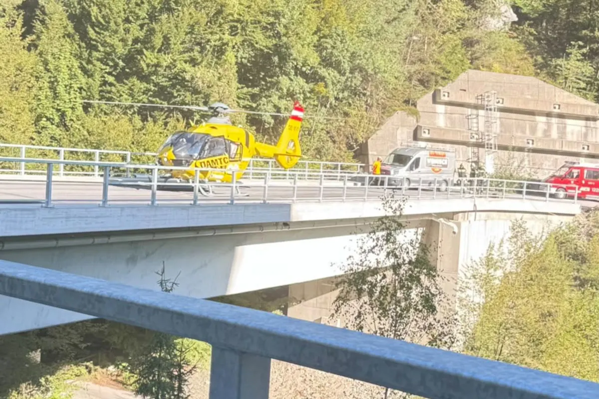 Ein gelber Rettungshubschrauber steht auf einer Brücke in einer bergigen Landschaft. Hinter ihm sind ein Einsatzfahrzeug und ein Kleintransporter zu sehen.