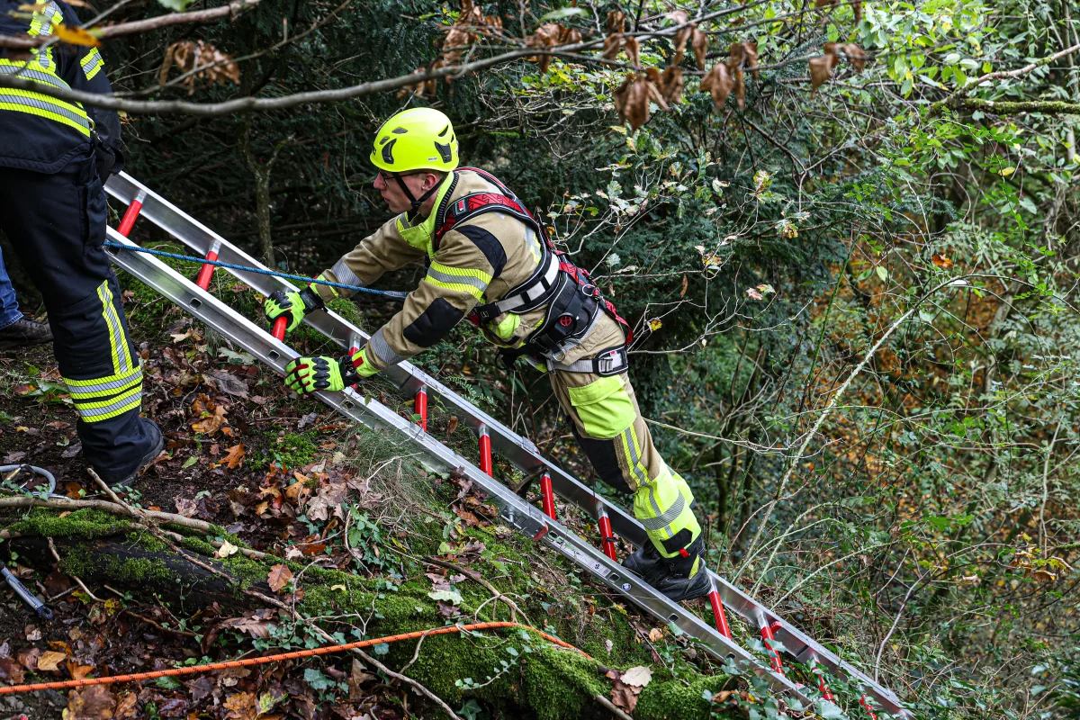 Ein Feuerwehrmann in leuchtender Ausrüstung steigt eine mit Seil gesicherte Leiter in einem steilen, bewaldeten Gelände hinauf.
