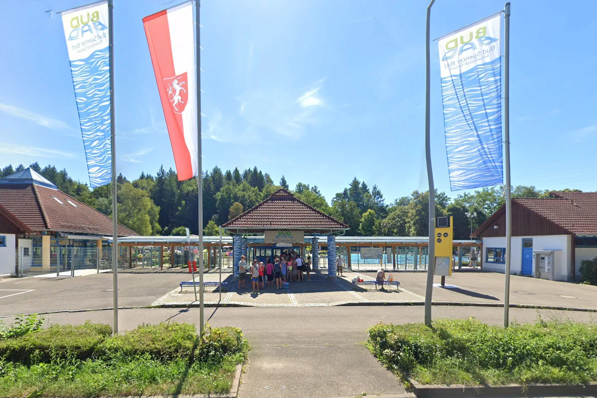Ein Schwimmbad-Eingang mit mehreren Personen davor, umgeben von Flaggen und Bäumen im Hintergrund.