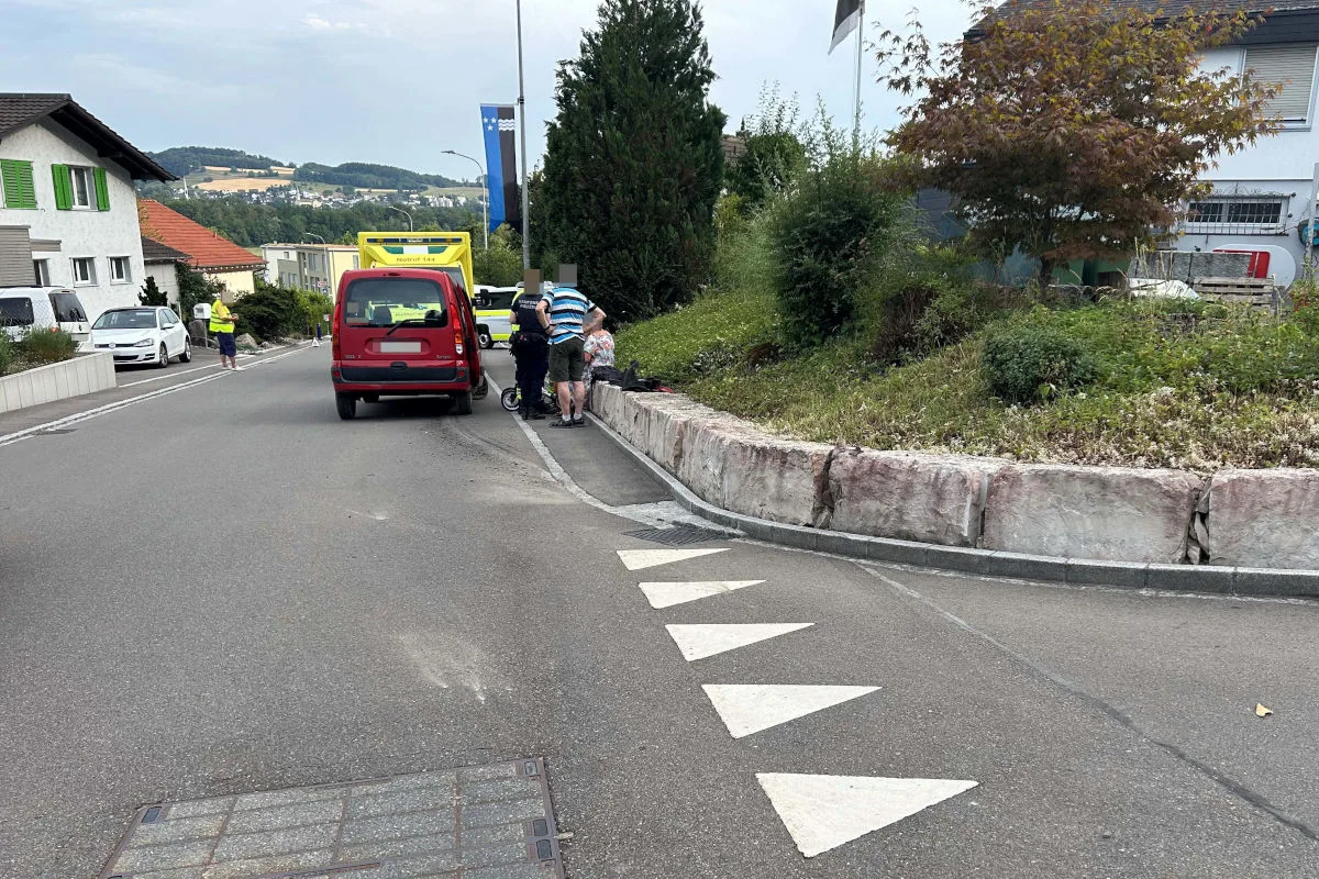 Ein rotes Auto steht auf einer Straße neben einer Gruppe von Menschen und einem gelben Krankenwagen. Das Umfeld wirkt ländlich.