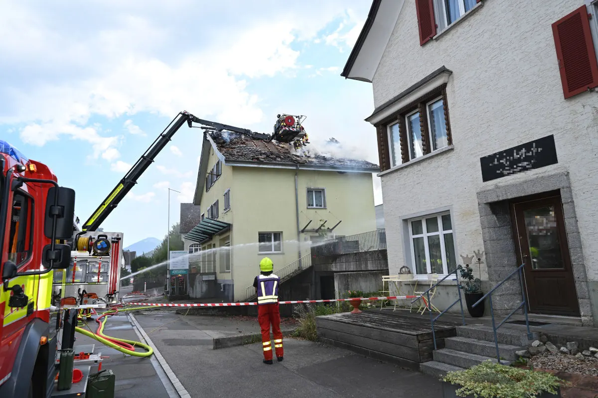 Ein Feuerwehrmann spritzt Wasser auf ein brennendes Dach eines Wohnhauses. Ein Feuerwehrauto und Absperrband sind sichtbar.