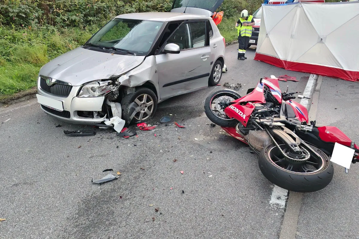 Ein silberner Skoda mit beschädigter Front steht neben einem umgekippten roten Honda-Motorrad auf einer Straße. Im Hintergrund sind zwei Personen in Warnwesten und eine Schutzwand zu sehen.