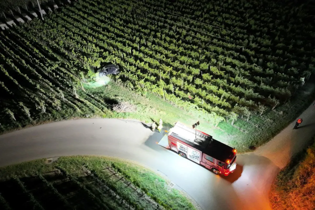 Ein Auto ist nachts in einen Weinberg gefahren. Ein Feuerwehrfahrzeug ist auf einer nahen Straße sichtbar.