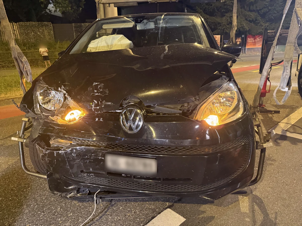 Ein frontal beschädigter VW mit eingeschalteten Scheinwerfern wird bei Nacht abgeschleppt. Im Hintergrund ist ein Zaun und eine Person sichtbar.