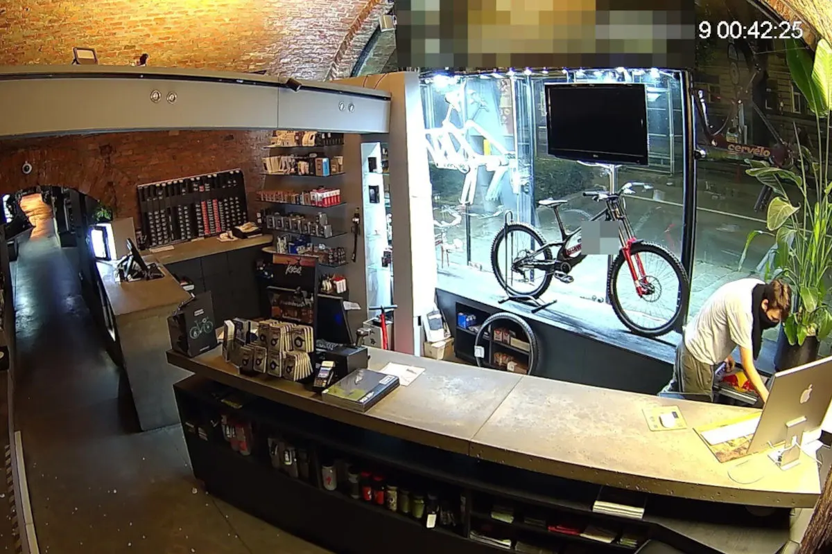 Eine Überwachungskameraaufnahme zeigt eine Person, die sich hinter dem Tresen eines Fahrradgeschäfts bückt. Im Hintergrund ist ein ausgestelltes Fahrrad im Schaufenster zu sehen.