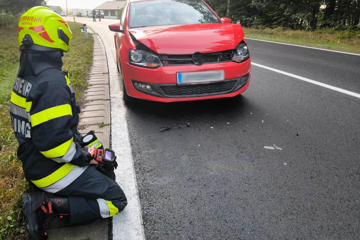 Ein Feuerwehrmann in Schutzkleidung kniet neben einem roten Auto mit beschädigter Front auf einer Landstraße. Im Hintergrund sind weitere Einsatzkräfte zu sehen.