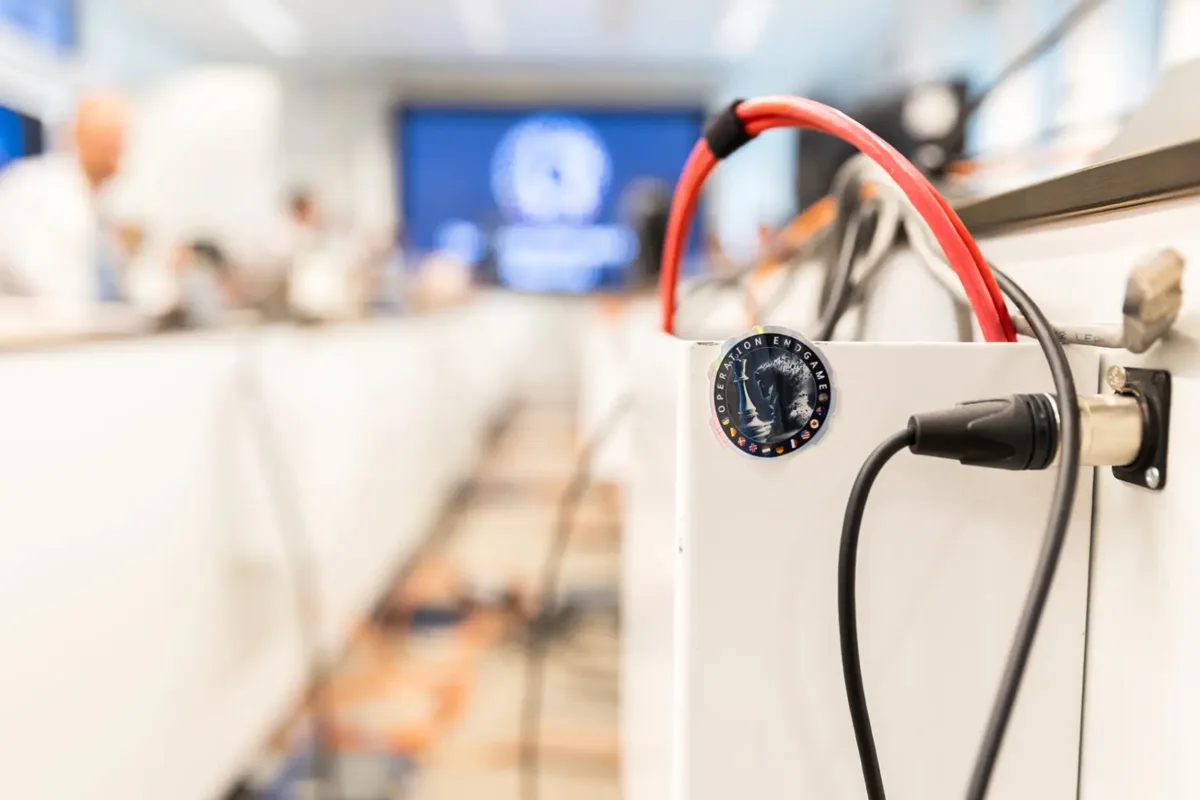 Ein weißes elektronisches Gerät mit verbundenen Kabeln. Ein Aufkleber mit der Aufschrift 'Operation Endgame' und mehreren Flaggen ist sichtbar.